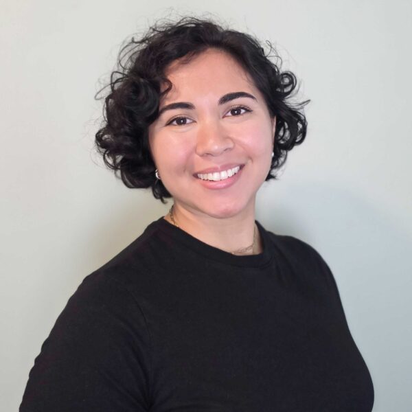 Tori Lipnicky - Woman with short curly hair wearing a black t-shirt smiling in front of a plain light-colored background.