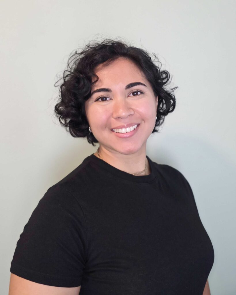 Tori Lipnicky - Woman with short curly hair wearing a black t-shirt smiling in front of a plain light-colored background.